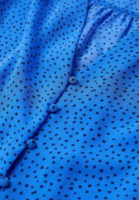 Blouse bleue transparente avec un petit motif noir moucheté, dotée d'un devant boutonné et d'un col froncé. Texture lisse et tissu léger.