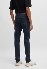 Dunkelblaue Denim-Jeans mit Slim-Fit, ausgestattet mit Gesäßtaschen und einem schwarzen Lederpatch mit BOSS-Logo. Stilvoll kombiniert mit einem schwarzen Hemd und weißen Schuhen.