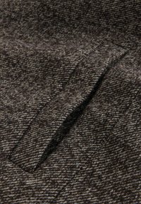 Vilnas audums tumši brūnā un bežā houndstooth rakstā. Bieza, teksturēta auduma struktūra ar redzamām šķiedrām. Satur diskrētu kabatu uz virsmas.