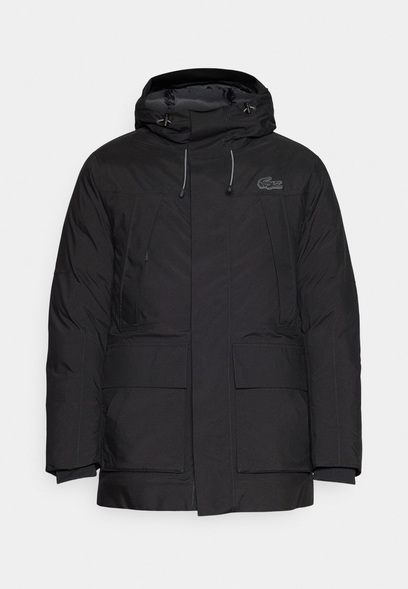 Chaqueta acolchada negra con cuello alto, capucha con cordón, dos grandes bolsillos delanteros y puños acanalados. Presenta un logo en la zona del pecho.