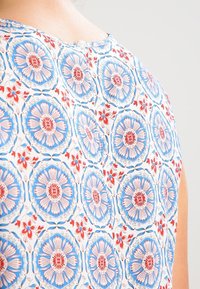 Top sans manches avec un motif floral composé de fleurs bleues, roses et rouges sur un fond blanc. Fermeture au dos avec un bouton.