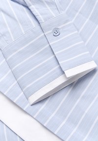 Camicia azzurro chiaro con strisce bianche, dotata di polsini con bottone e orlo bianco. Il tessuto appare liscio e leggermente strutturato.