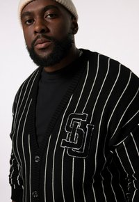 Zwart-witte gestreepte cardigan met een V-hals, knoopsluiting en een logo-applicatie. De stof lijkt gebreid; textuur met ribboorden en zoom.