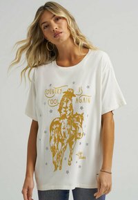 Hvid bomulds t-shirt med et vintage cowboy design i guld på hesteryg, med sølvstjernes detaljer og teksten "Country is Cool Again."