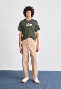 Oliven grøn kortærmet Nike t-shirt med hvidt og lime logo, parret med beige brede bukser og hvide sneakers med gummisåler.