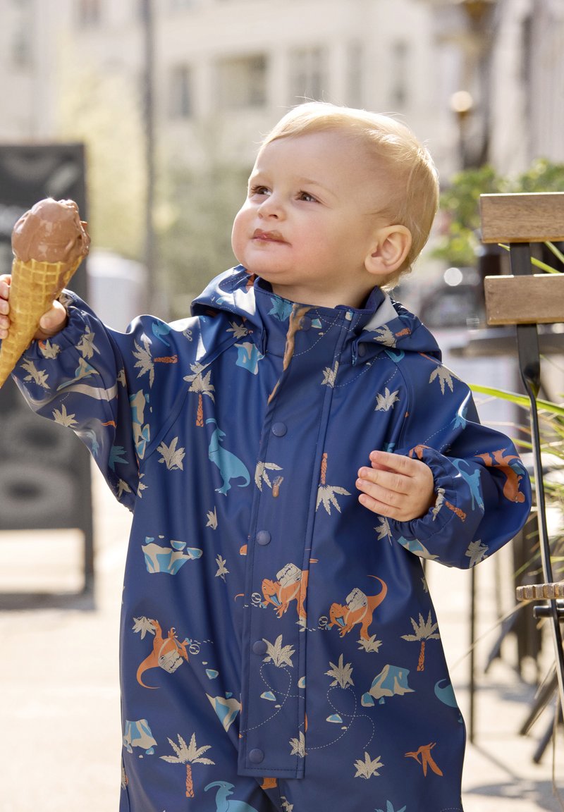 Blauwe waterdichte regenjas met dinosaurusthema in oranje, grijs en teal. Bevat een capuchon en drukknoopsluitingen, vastgehouden door een kind met een ijsje.