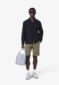 Chemise bleu marine à boutons, shorts olive, baskets grises et un sac bleu clair. Le mannequin porte des lunettes de soleil ; design minimal avec un logo discret.