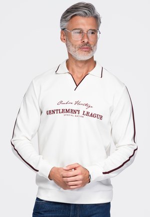 Homme aux cheveux gris et à la barbe, portant des lunettes transparentes et une chemise blanche à manches longues avec une bordure bordeaux et le texte "Gentlemen's League".