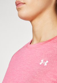 T-shirt desportivo rosa claro, feita de tecido suave. Apresenta um decote redondo e um logótipo branco da Under Armour no peito. Textura suave.