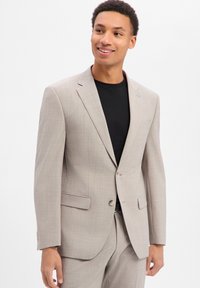 Junger Mann mit Lächeln, trägt eine hellbeige Anzugjacke und passende Hose über einem schwarzen Rundhalsausschnitt, steht vor einem weißen Hintergrund.