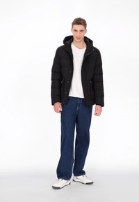 Černá péřová bunda s kapucí, se zipem na přední straně a elastickými manžetami, v kombinaci s modrými širokými džínami a bílými teniskami.