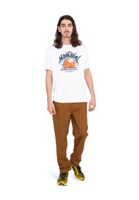 Vit t-shirt med en färgglad grafik som står "Montréal", ihop med bruna byxor och svarta och gula sneakers.