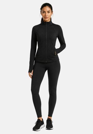 Frau steht und trägt eine enganliegende schwarze Reißverschlussjacke, passende schwarze Leggings und schwarze Sportschuhe vor einem schlichten hellen Hintergrund.