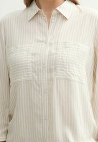 Light beige button-up shirt with vertical white stripes, two front pockets, and a standard collar. Made from a soft, textured fabric.