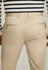 Pantaloni beige con una tessitura liscia, taglio dritto e tasche posteriori. Indossati con un maglione a righe blu navy e crema.