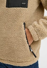 Beige fleece pullover with a textured, cozy surface, featuring a black zippered chest pocket and side pockets with contrasting zipper branding.