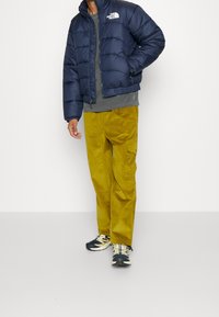 Marineblå puffjacka med quiltad struktur och frontdragkedja, grå T-shirt, senapsgula kordfläktbyxor och svarta skor med vita sulor.