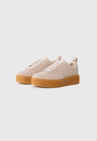 Beige semskede lavtopp sneakers med tykke ribbet gummisåler, hvite snørebånd, hanker og liten gull "SOREL" branding på siden.
