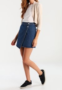 Denimová sukně s křížovým designem, světle modré barvy, s knoflíkovými detaily, v kombinaci s bílým topem a béžovým kardigánem, černými teniskami.