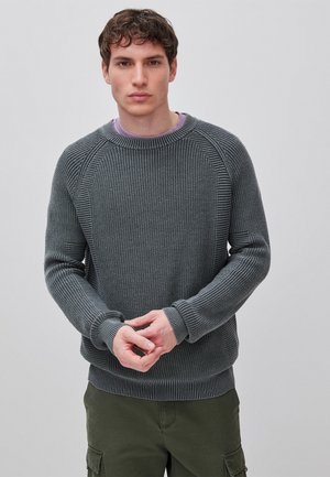 Giovane uomo con capelli castani ricci, indossa un maglione grigio a maglia e pantaloni cargo verde oliva, in piedi davanti a uno sfondo chiaro e uniforme.