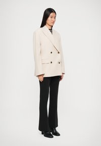 Woman standing wearing oversized cream double-breasted blazer, black flared pants, black ankle boots, and a black and white striped top.