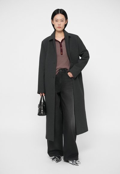 Eine Frau trägt einen langen dunkelgrauen Mantel, weite schwarze Jeans, ein braunes Hemd mit Knöpfen, silberne spitze Schuhe und hält eine kleine schwarze Handtasche.