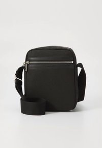 Juoda per petį nešiojama rankinė, pagaminta iš tvaraus medžiagos, su priekinė kišene su užtrauktuku, reguliuojama dirželiu ir stačiakampio formos minimalistiniu dizainu.