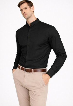 Homme portant une chemise noire à col mao rentrée dans un pantalon beige avec une ceinture marron, les mains dans les poches, sur fond blanc.