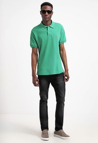 Grön pikétröja i bomull, svarta slim-fit jeans och grå sneakers. Modell med mörka solglasögon. Enkel design utan mönster.