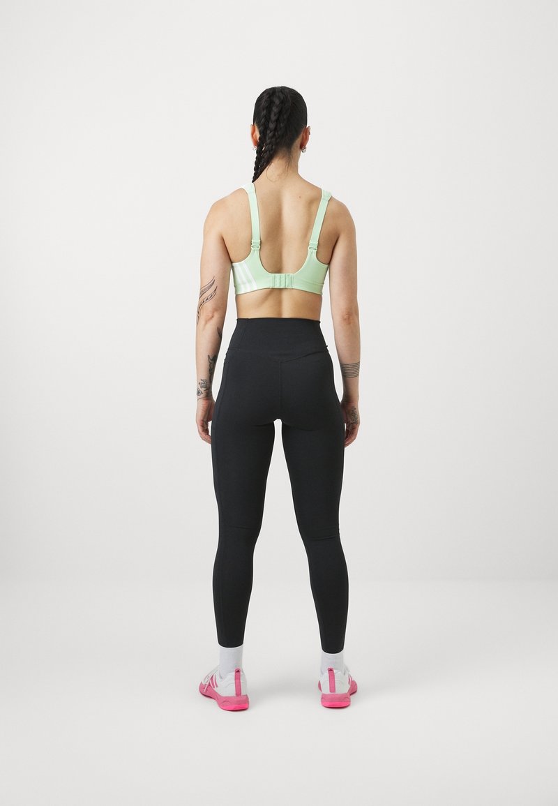 Grön sportbh med justerbara band, i kombination med svarta högmidjade tights och rosa sportskor. Slät textur, minimalistisk design.