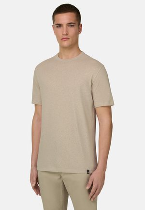 Jeune homme aux cheveux courts portant un T-shirt beige à manches courtes et un pantalon assorti, debout devant un fond clair uni.