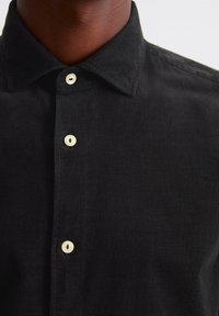 Camisa de pana negra con cuello puntiagudo, que presenta botones blancos y una sutil textura de ribete en toda la tela.