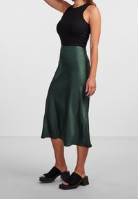 Falda midi de satén verde con un diseño fluido, combinada con una camiseta negra de canalé y zapatos de plataforma negros. Textura suave, brillo sutil.