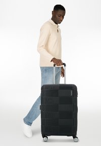 Maleta rígida negra con diseño de cuadrícula texturizada, equipada con un mango retráctil y cuatro ruedas para mayor movilidad.