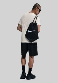Mochila negra con cordón y logo blanco de Nike, llevada en la espalda de una persona. La persona viste una camiseta beige clara y pantalones cortos negros, con zapatillas negras.