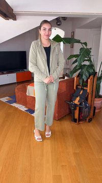 Νεαρή γυναίκα με ελαφρύ μπουφάν και παντελόνι στέκεται σε ξύλινο πάτωμα σε καθιστικό με καναπέ, κωπηλατική μηχανή, φυτά και τηλεόραση στο βάθος.