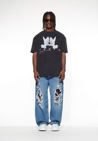 Schwarzes übergroßes T-Shirt mit weißem Vogel-Graphic, kombiniert mit ausgebleichten blauen Jeans mit großen Rissen. Weiße Sneakers runden den Look ab.