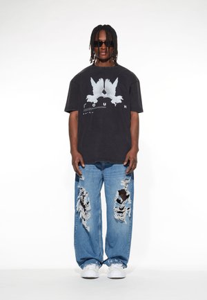Sort oversized t-shirt med hvid fugle-grafik, parret med slidte blå jeans med store huller. Hvide sneakers fuldender Looket.