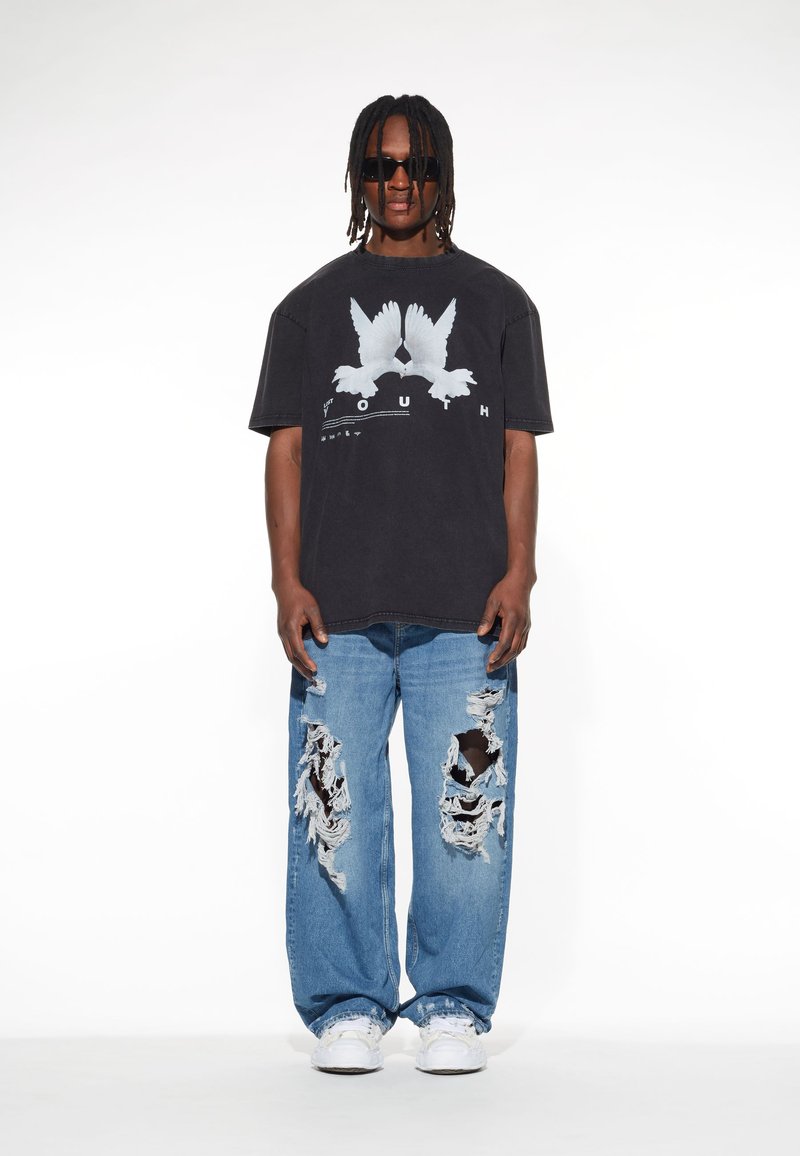 Schwarzes übergroßes T-Shirt mit weißem Vogel-Graphic, kombiniert mit ausgebleichten blauen Jeans mit großen Rissen. Weiße Sneakers runden den Look ab.