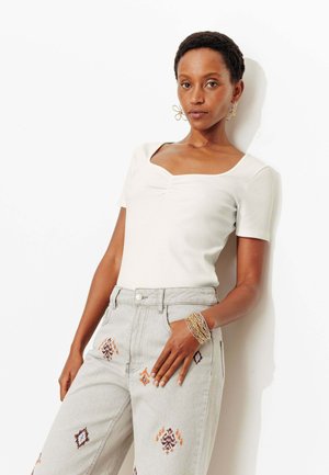 Femme aux cheveux courts portant un haut blanc plissé, un pantalon taille haute gris clair avec des motifs brodés, des boucles d'oreilles dorées et des bracelets perlés.