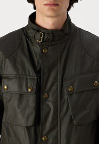 Veste légère de couleur vert foncé avec un col haut, des boutons-pression en laiton et deux poches poitrine. Texture lisse, design minimaliste.