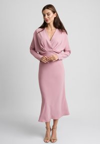 Długa różowa sukienka z dekoltem w serek i długimi rękawami, z zebranym stanikiem i zwiewną spódnicą. Gładka faktura i elegancka sylwetka.