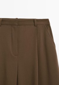 Pantaloni marroni su misura con una texture liscia, dotati di tasche laterali e design a gamba dritta. Dettagli di cucitura minimi vicino alle tasche.