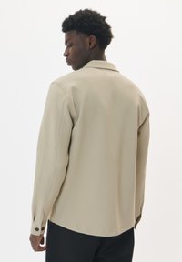 Camicia beige a maniche lunghe con una consistenza morbida, caratterizzata da un colletto all'italiana e polsini con bottoni. Il retro presenta un design semplice e liscio senza motivi.