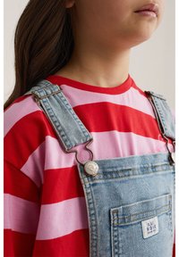 Dítě v červeném a růžovém pruhovaném tričku pod světle modrými džínovými laclovými kalhotami s kovovými zapínáními a přední kapsou s nápisem "Blue Ridge".