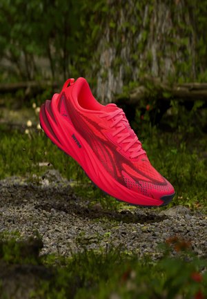 Scarpa da ginnastica rosso brillante con suola spessa che fluttua sopra ghiaia e terreno verde coperto di muschio in un ambiente naturale all'aperto.