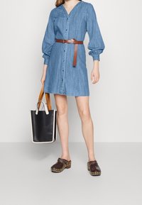 Jeansowa sukienka koszulowa z dekoltem w serek, długimi bufkowymi rękawami, brązowym skórzanym paskiem, zestawiona z brązowymi klapkami z ćwiekami oraz torbą typu tote w czarno-białe wzory.