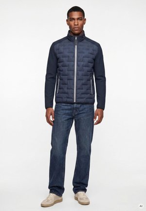 Mann trägt eine marineblaue gesteppte Jacke mit Reißverschluss, blaue Jeans und beige Sneakers, steht vor einem schlichten weißen Hintergrund.