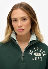 Maglione verde con zip e ricamo con la scritta "SO. TRACK DEPT 86". Sotto si intravede una camicia con colletto a righe blu e bianche, visibile dal colletto.