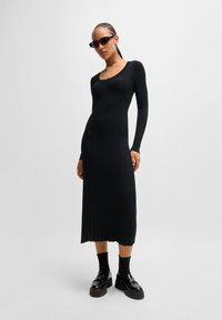 Black ribbed long-sleeve dress with a scoop neckline, fitted silhouette, and a knee-length hem. Model wears chunky black platform shoes and sunglasses.